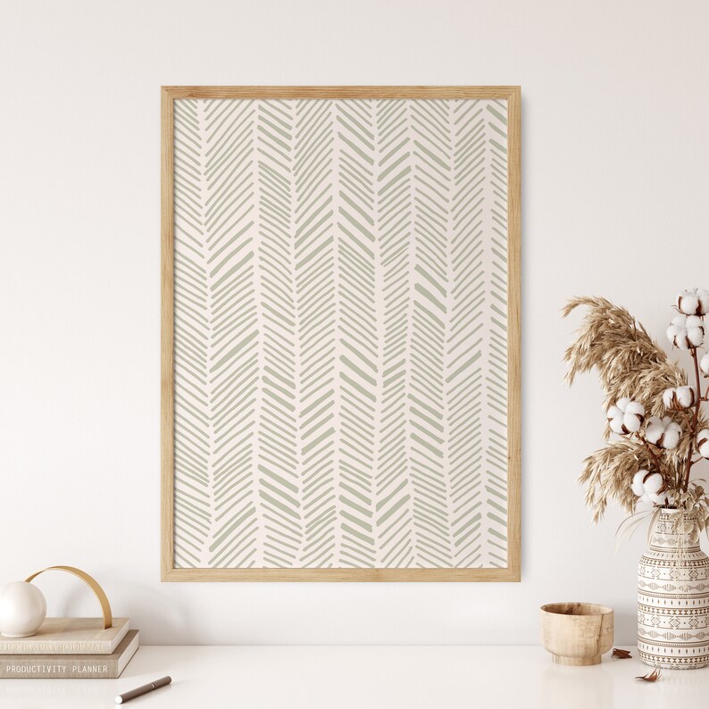 May include: A framed print with a beige and white chevron pattern. The print is hanging on a white wall above a desk with a book, a pencil, and a vase with dried flowers.