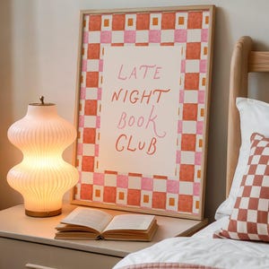 May include: A framed print with the words "LATE NIGHT BOOK CLUB" in pink script, set against a white background and a border of orange and pink checks. A lit, ribbed glass lamp and an open book sit on a bedside table. A checkered pillow is on the bed.