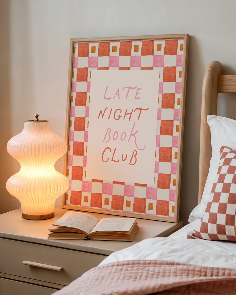 May include: A framed print with the words "LATE NIGHT BOOK CLUB" in pink script, set against a white background and a border of orange and pink checks. A lit, ribbed glass lamp and an open book sit on a bedside table. A checkered pillow is on the bed.