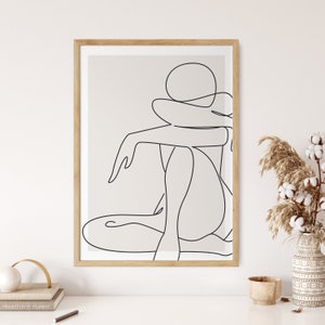 Op de afbeelding: Een lijntekening van een persoon die zit met de armen om het lichaam gewikkeld. De tekening is in zwart-wit en is ingelijst in een licht houten lijst.