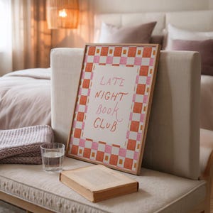 May include: A framed print with the text "LATE NIGHT BOOK CLUB" in pink script. The frame has a checkerboard border in pink and orange. A book and a glass of water sit on a bench next to the print.