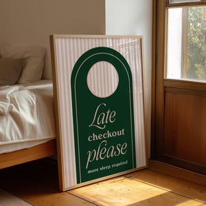 Op de afbeelding: Ingelijste wandkunst met een groen deurhangerontwerp en de woorden "Late checkout please". De achtergrond heeft verticale roze en witte strepen. Het frame heeft een lichte houtkleur. De tekst "more sleep required" is ook zichtbaar.