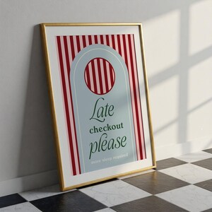 Op de afbeelding: Een ingelijste kunstdruk met een gouden rand. Het kunstwerk toont een lichtblauwe deurhanger vorm met de woorden "Late checkout please" in groen schrift. De achtergrond heeft rode en witte verticale strepen.