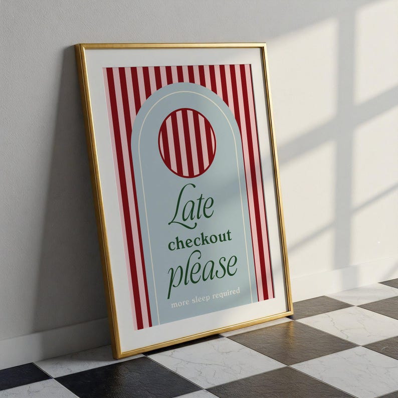 Op de afbeelding: Een ingelijste kunstdruk met een gouden rand. Het kunstwerk toont een lichtblauwe deurhanger vorm met de woorden "Late checkout please" in groen schrift. De achtergrond heeft rode en witte verticale strepen.