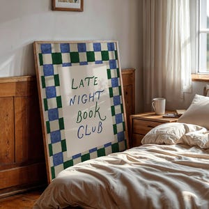 May include: A framed poster with the text "LATE NIGHT BOOK CLUB" in a bedroom. The poster has a green and blue checkered border. The room includes a bed with beige bedding, a wooden bedside table, and a window with a white curtain.