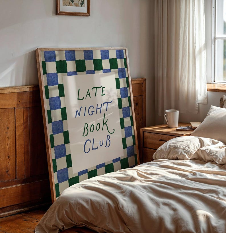 May include: A framed poster with the text "LATE NIGHT BOOK CLUB" in a bedroom. The poster has a green and blue checkered border. The room includes a bed with beige bedding, a wooden bedside table, and a window with a white curtain.