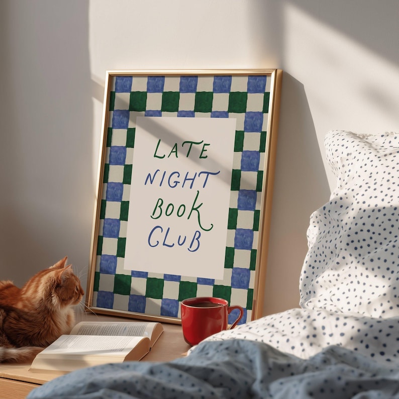May include: A framed print with the text "LATE NIGHT BOOK CLUB" in green and blue script. The frame has a blue, green, and white checkered border. A red mug, an open book, and a ginger cat are on a wooden surface.