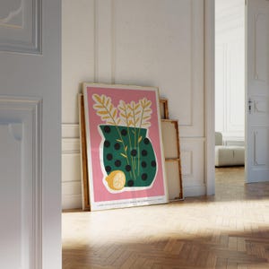 May include: A green vase with black polka dots filled with yellow wheat stalks and a lemon slice on a pink background. The vase is sitting on a table with a wooden floor.