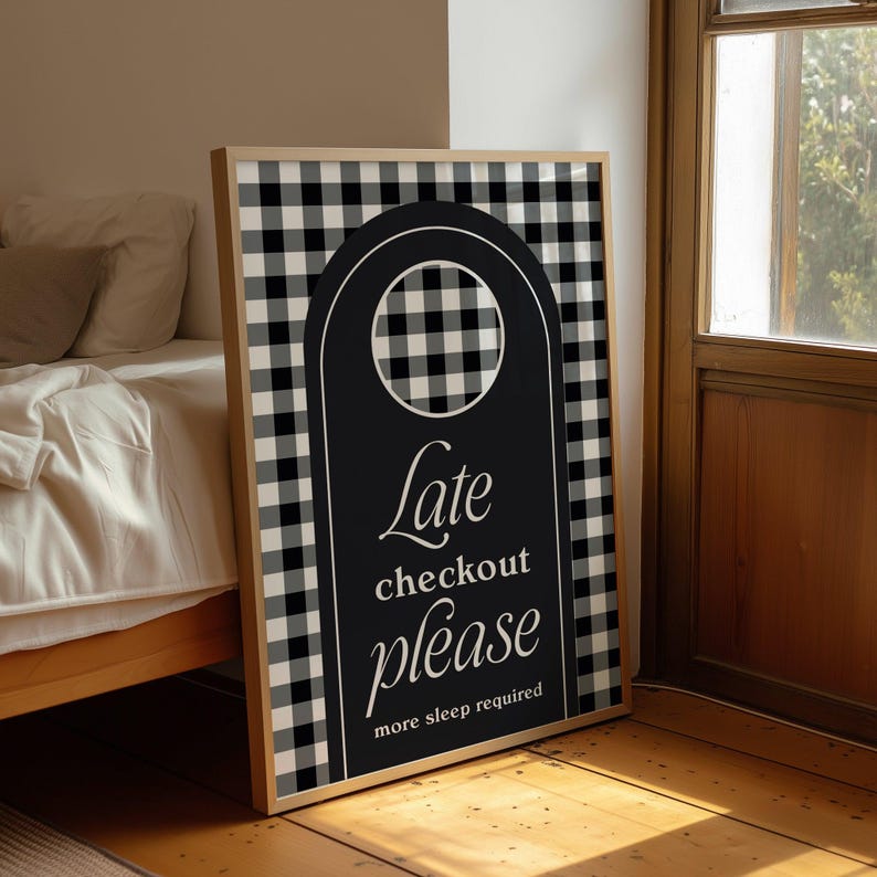 Op de afbeelding: Een zwart-wit geruite print in een lijst met een zwart deurhangerontwerp. De deurhanger leest "Late checkout please more sleep required" in wit script. De lijst heeft een lichte houtkleur.