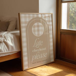 Op de afbeelding: Een ingelijst bord met een beige en wit geruit patroon en de woorden "Late checkout please more sleep required" in een scriptlettertype. Het bord heeft een licht houten frame en leunt tegen een houten vloer.