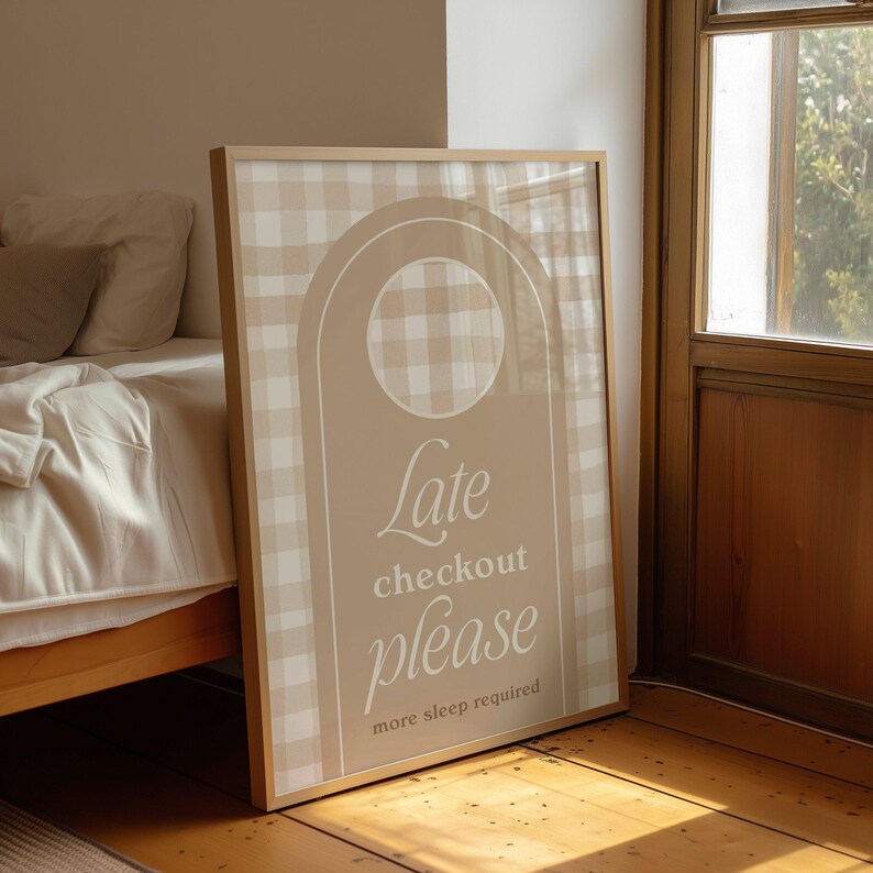 Op de afbeelding: Een ingelijst bord met een beige en wit geruit patroon en de woorden "Late checkout please more sleep required" in een scriptlettertype. Het bord heeft een licht houten frame en leunt tegen een houten vloer.