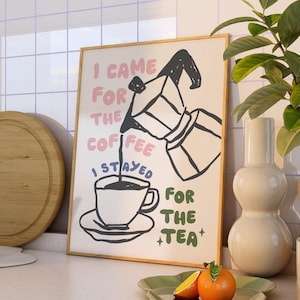 May include: A framed print with the text "I came for the coffee I stayed for the tea". The illustration shows a coffee pot pouring into a cup. A wooden chopping board and a plant are in the background. The print is in a kitchen setting.