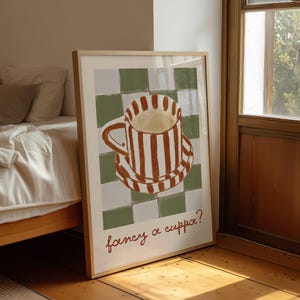 May include: A framed art print featuring a whimsical illustration of a striped coffee cup and saucer. The cup and saucer are brown and white striped, set against a green and white checkered background. The text "fancy a cuppa?" is written in brown script.