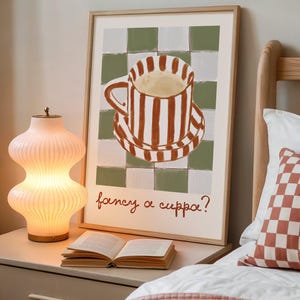 May include: A framed art print featuring a striped teacup and saucer on a checkered background, with the text "fancy a cuppa?" in red script. A lit, ribbed glass lamp and an open book sit on a nightstand.