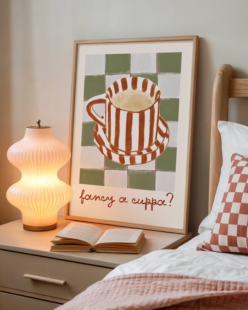 May include: A framed art print featuring a striped teacup and saucer on a checkered background, with the text "fancy a cuppa?" in red script. A lit, ribbed glass lamp and an open book sit on a nightstand.