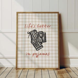Pu&ograve; includere: Una stampa incorniciata con uno sfondo a quadretti beige e bianco. La stampa presenta uno schizzo di pigiama e il testo "life's better in pyjamas" in corsivo. La cornice &egrave; in legno.