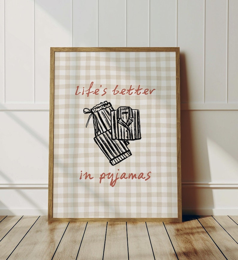 Pu&ograve; includere: Una stampa incorniciata con uno sfondo a quadretti beige e bianco. La stampa presenta uno schizzo di pigiama e il testo "life's better in pyjamas" in corsivo. La cornice &egrave; in legno.