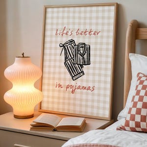 Pu&ograve; includere: Una stampa incorniciata con uno sfondo a quadretti beige e bianco e la scritta "life's better in pyjamas" in rosso. La stampa presenta un'illustrazione in bianco e nero di un pigiama. Una lampada in vetro rosa a coste illuminata e un libro aperto sono su un comodino.