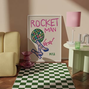 May include: A pink and green chequered rug with a pink lamp, a white side table, and a framed poster with the text "ROCKET MAN 1972" and a drawing of a disco ball with legs wearing star-shaped shoes.