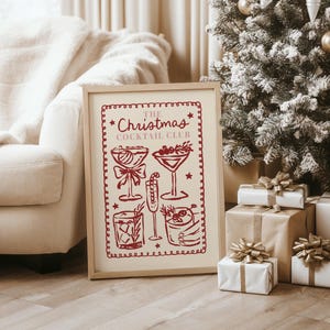 Op de afbeelding: Een ingelijste print met de tekst "The Christmas Cocktail Club" in het rood. De print bevat illustraties van verschillende cocktails. De lijst is lichtbruin en de print leunt tegen een witte bank en een kerstboom.