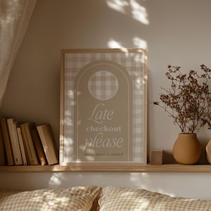 Op de afbeelding: Een ingelijste print met een beige en wit geruit patroon en de woorden "Late checkout please more sleep required". De print staat op een houten plank met boeken, een vaas met droogbloemen en een klein houten blok.