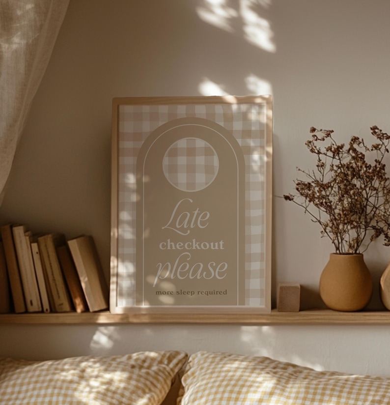 Op de afbeelding: Een ingelijste print met een beige en wit geruit patroon en de woorden "Late checkout please more sleep required". De print staat op een houten plank met boeken, een vaas met droogbloemen en een klein houten blok.