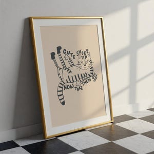 Op de afbeelding: Ingelijste kunstprint met een illustratie van een kat met de tekst "Cute Shoes Take Them Off Now". De kat is zwart en wit, liggend op een beige achtergrond, omlijst door een gouden lijst.