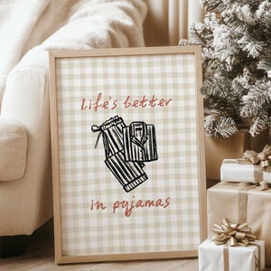 Pu&ograve; includere: Una stampa incorniciata con uno sfondo a scacchi beige e bianco. La stampa presenta un'illustrazione in bianco e nero di un pigiama e il testo "life's better in pyjamas" in corsivo rosso. La cornice &egrave; marrone chiaro. L'immagine &egrave; accanto a un albero di Natale e regali confezionati.
