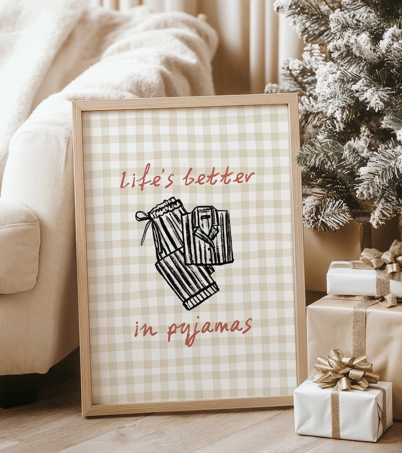 Pu&ograve; includere: Una stampa incorniciata con uno sfondo a scacchi beige e bianco. La stampa presenta un'illustrazione in bianco e nero di un pigiama e il testo "life's better in pyjamas" in corsivo rosso. La cornice &egrave; marrone chiaro. L'immagine &egrave; accanto a un albero di Natale e regali confezionati.