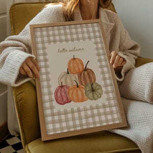 May include: A framed print with a watercolour illustration of pumpkins in shades of orange, brown, pink, white, and green. The print has a beige and white checked border and the words "hello autumn" in a script font.