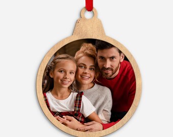 Könnte beinhalten: Weihnachtsbaumschmuck aus Holz in Form einer Kugel mit einem Familienporträt im Inneren. Der Schmuck ist aus hellem Holz gefertigt und hat ein rotes Band zum Aufhängen. Die Familie besteht aus einem Mädchen, einer Frau und einem Mann, die alle lächeln.