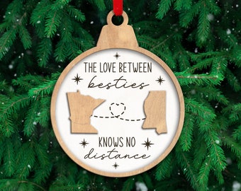 May include: A round wooden ornament with a red ribbon hanger featuring a white center insert. The text reads THE LOVE BETWEEN besties KNOWS NO distance. Two wooden state silhouettes are connected by a dashed line forming a heart shape.