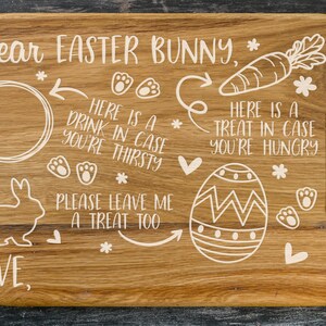 May include: Wooden cutting board with a white design of an Easter bunny, a carrot, an egg, and paw prints. The board has a handle and the text "Dear Easter Bunny, * LOVE * HERE IS A DRINK IN CASE YOU'RE THIRSTY * PLEASE LEAVE ME A TREAT TOO * HERE IS A TREAT IN CASE YOU'RE HUNGRY *" is written on it.
