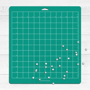 May include: A green cutting mat with a grid pattern and white numbers. There are small, white, diamond-shaped rhinestones scattered on the mat and on a white surface below the mat.