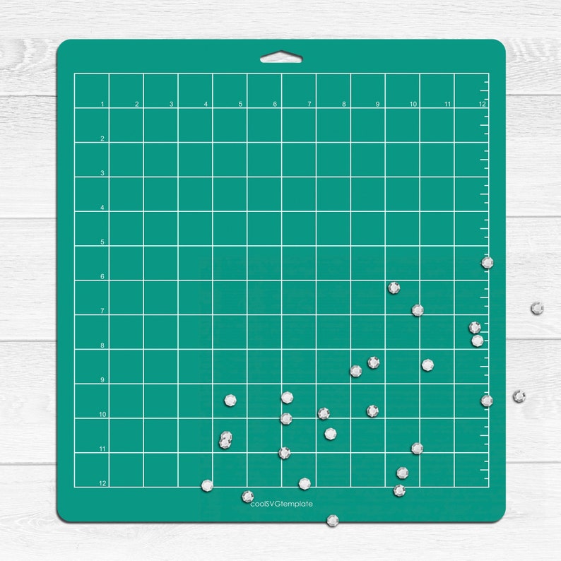 May include: A green cutting mat with a grid pattern and white numbers. There are small, white, diamond-shaped rhinestones scattered on the mat and on a white surface below the mat.