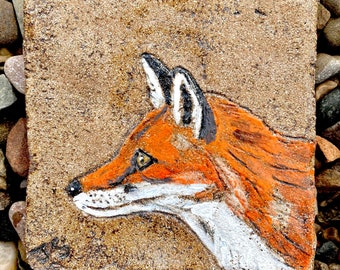 May include: A close-up of a hand-painted ceramic tile featuring a fox's head. The fox is painted in shades of orange, white, and black, with a detailed depiction of its fur and eyes.