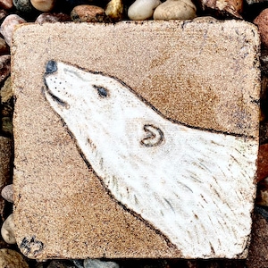 Peut inclure: Un carreau de céramique blanc avec une image peinte à la main d'une tête d'ours polaire regardant vers le haut. Le carreau a un fond brun et une texture rugueuse.