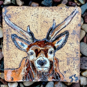 May include: A hand-painted ceramic tile depicting a brown and white deer with antlers. The tile has a rustic, textured surface and a light brown background.