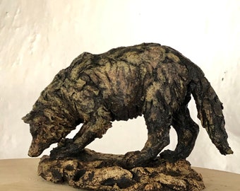 May include: A ceramic sculpture of a wolf, standing on a rocky base. The wolf is painted in shades of brown and green, with a textured surface that resembles bark.