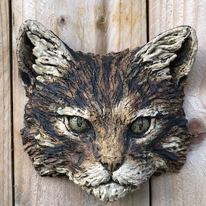 May include: A ceramic cat head wall sculpture with realistic details. The cat has brown, black, and white fur with green eyes. The sculpture is mounted on a wooden surface.