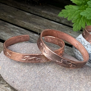 May include: Copper cuff bracelets with a running hare design. The bracelets are a warm reddish-brown color and rest on a gray stone. The design is etched into the metal, adding texture and detail to the jewelry.