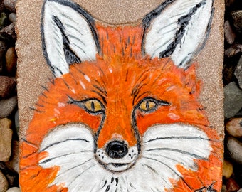 May include: A close-up of a ceramic tile with a hand-painted orange fox with white fur and black eyes. The fox's face is the main focus of the image.