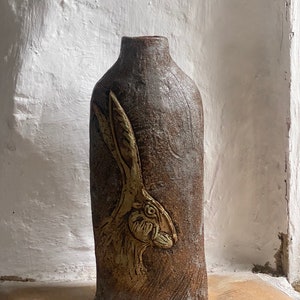 Op de afbeelding: Een bruine keramische vaas met een getextureerd oppervlak en een reliëfdesign van een hazenkop in goud.