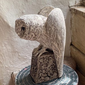 Op de afbeelding: Een gedetailleerde, gebroken witte uil sculptuur met getextureerde vleugels en veren, geplaatst op een vierkante, gebeeldhouwde basis. De kop van de uil is gedraaid en zijn ogen zijn zichtbaar. De sculptuur staat op een donkergroen, cirkelvormig oppervlak.
