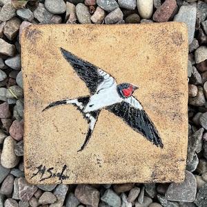 May include: A ceramic tile featuring a painted image of a black and white swallow with a red beak. The tile is beige and has the signature "M.S." in the bottom left corner.