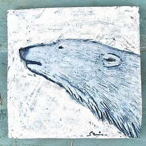 May include: A white ceramic tile with a blue and white relief sculpture of a polar bear's head. The bear's head is facing to the left and the sculpture is set against a white background.