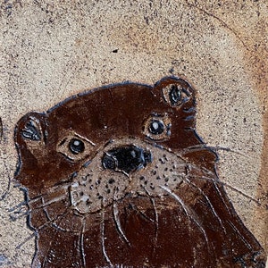 Puede incluir: Primer plano de una nutria de cerámica marrón con una superficie texturizada. La nutria tiene una expresión juguetona con la boca ligeramente abierta y sus bigotes visibles.