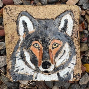 May include: A square, hand-painted ceramic tile depicting a wolf's face. The wolf has black and white fur, orange accents around the eyes and nose, and yellow eyes. The tile has a textured, tan background and is surrounded by small stones.