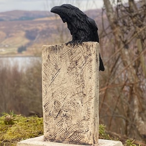 May include: A black ceramic raven sculpture perched on a white ceramic pedestal with a textured surface. The pedestal has a square base. The sculpture is set against a blurred background of a landscape with trees and a body of water.