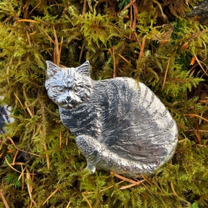 May include: A silver-colored pewter cat figurine, sitting with its head turned to the side, is displayed on a bed of green moss.